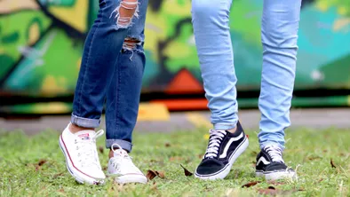 Ноги подростков в рваных джинсах и кедах на фоне травы и граффити на стене