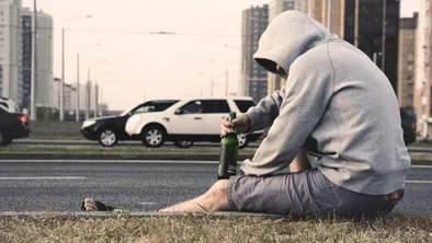 Мужчина с пивом в толстовке и шортах сидит на обочине автомобильной дороги